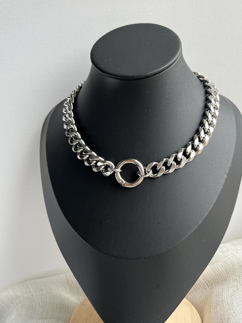 Op de afbeelding: Een zilverkleurige ketting met een grof geschakeld ontwerp. De ketting heeft een cirkelvormige sluiting in het midden en wordt gepresenteerd op een zwarte mannequin. Een opvallend sieraad.