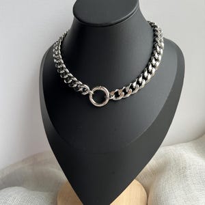 May include: A silver chain necklace with a chunky curb chain design. The necklace is displayed on a black mannequin bust. The necklace has a circular clasp in the centre.