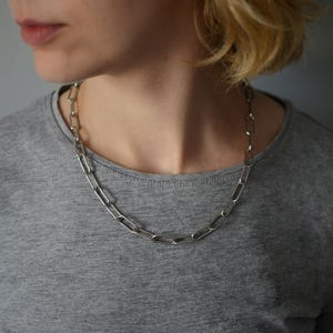 Puede incluir: Un collar de cadena en tono plateado con eslabones rectangulares alargados conectados en un patrón repetitivo. El collar descansa alrededor del cuello, mostrando un acabado metálico y un diseño estructurado compuesto por eslabones de metal abiertos y uniformes.