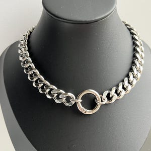 Op de afbeelding: Een zilverkleurige ketting met een chunky curb link design. De ketting heeft een cirkelvormige sluiting aan de voorkant. De sieraden worden gepresenteerd op een zwarte mannequin.