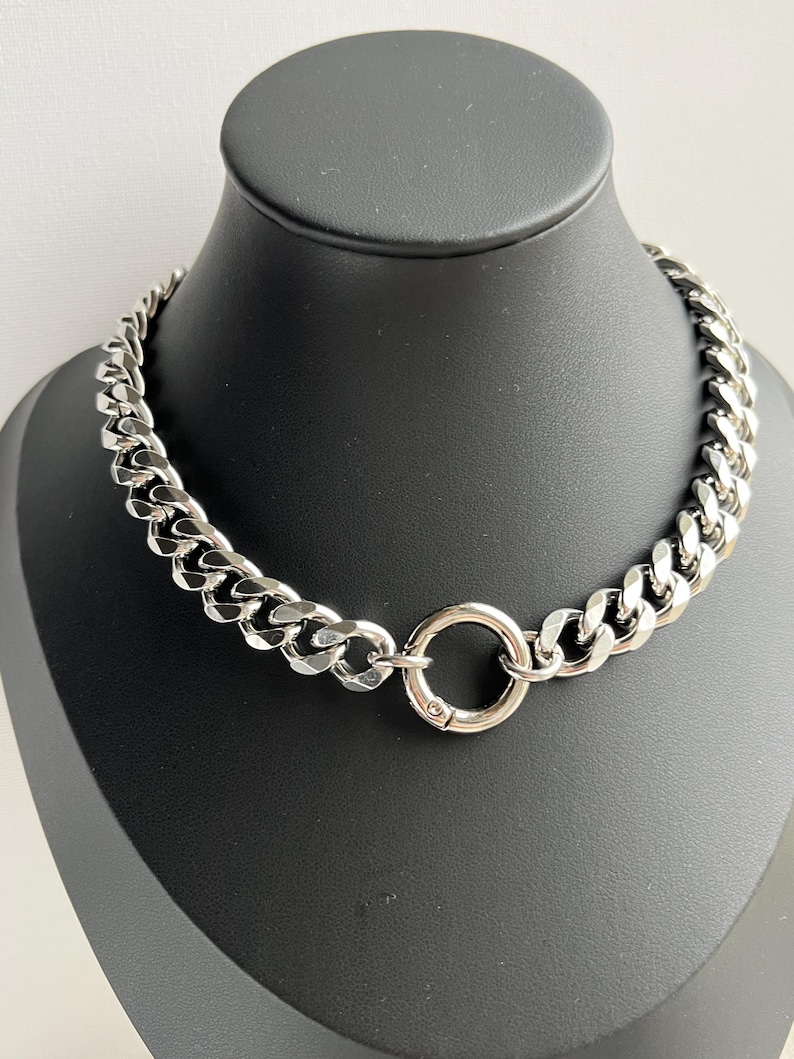 Op de afbeelding: Een zilverkleurige ketting met een chunky curb link design. De ketting heeft een cirkelvormige sluiting aan de voorkant. De sieraden worden gepresenteerd op een zwarte mannequin.