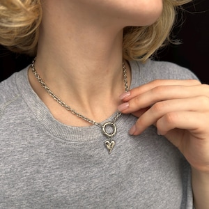 May include: Silver chain necklace with a heart pendant. The necklace has a chunky link chain and a small heart-shaped charm suspended from a circular clasp. The necklace is worn by a person wearing a grey top.
