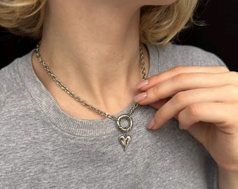 May include: Silver-toned chain necklace with a heart pendant. The necklace features a large link chain and a small heart-shaped charm hanging from a circular clasp. The necklace is worn on a person wearing a gray shirt.