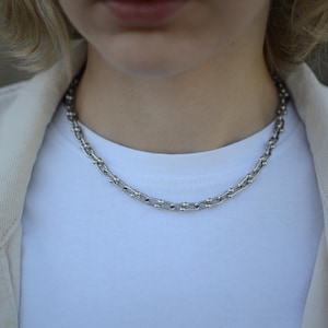 Peut inclure: Un collier à chaîne argentée composé d'une série de maillons rectangulaires allongés reliés par de petites perles sphériques polies à chaque jonction, créant un aspect texturé et métallique autour du cou.