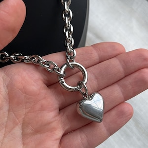 May include: A silver-toned chain necklace featuring a thick, polished link design. The necklace includes a circular spring-ring clasp that serves as a focal point, from which a smooth, puffed heart-shaped pendant hangs in a matching metallic finish.