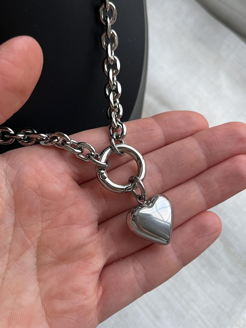 May include: A silver-toned chain necklace featuring a thick, polished link design. The necklace includes a circular spring-ring clasp that serves as a focal point, from which a smooth, puffed heart-shaped pendant hangs in a matching metallic finish.