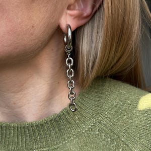 May include: Silver hoop earrings with a chain link dangle. The earrings are made of a shiny, metallic material and hang from the earlobe. The chain links are connected to each other and to the hoop, creating a stylish and modern look.