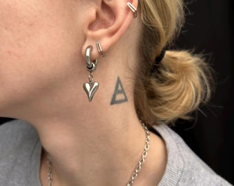 Op de afbeelding: Zilveren oorringen met een hartvormige hanger. De afbeelding toont meerdere oorbellen, waaronder een kleine staaf en een dubbele oormanchet. Een zilveren ketting is ook zichtbaar.