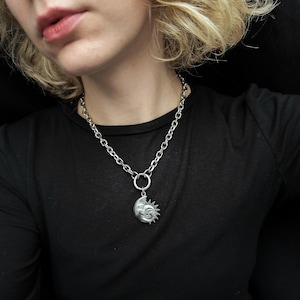 May include: Silver-toned necklace featuring a pendant with a smiling sun and moon design. The pendant hangs from a large link chain. The necklace is worn against a black top.