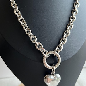 May include: A silver-toned necklace featuring a thick, chunky chain with large oval links. The necklace includes a circular spring-ring clasp at the centre, from which a smooth, polished, puffed heart-shaped pendant hangs as a focal point.