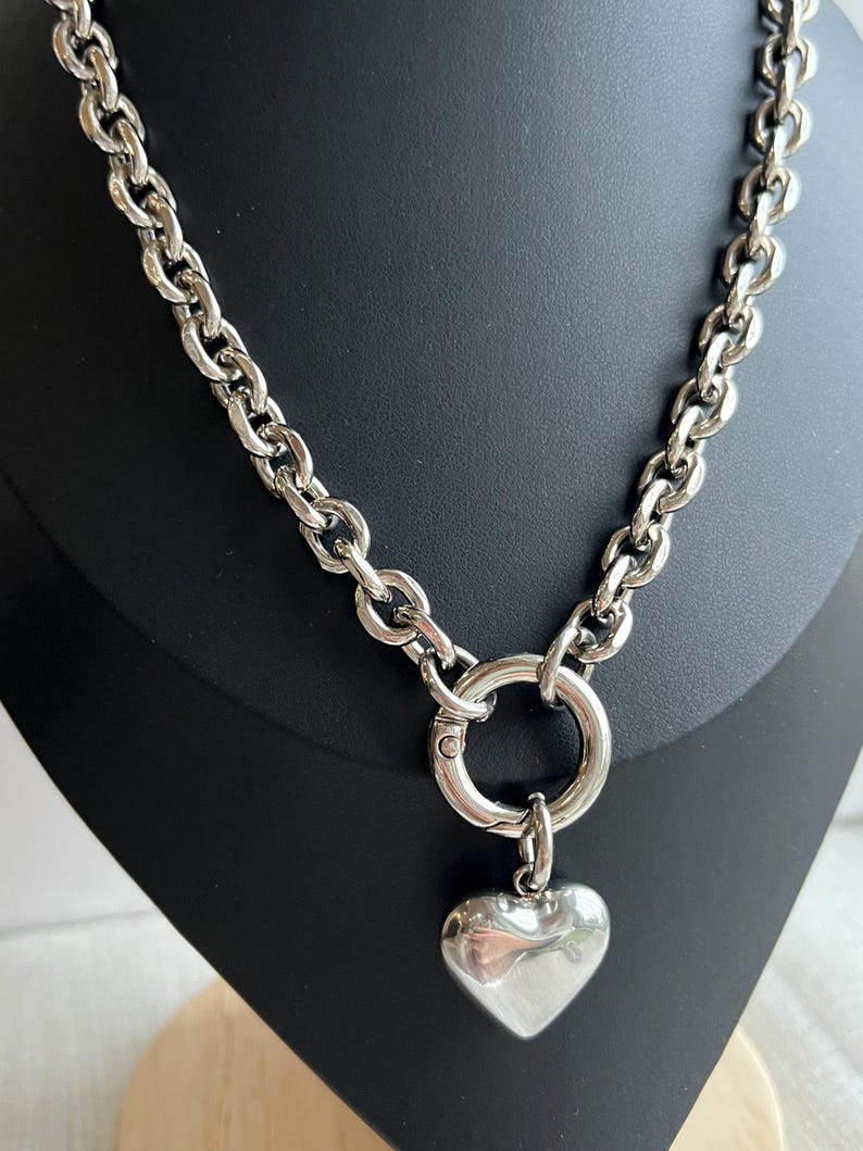 May include: A silver-toned necklace featuring a thick, chunky chain with large oval links. The necklace includes a circular spring-ring clasp at the centre, from which a smooth, polished, puffed heart-shaped pendant hangs as a focal point.