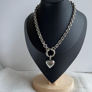 May include: A silver-colored necklace with a large heart pendant. The necklace features a chunky chain and a circular clasp. The heart pendant is smooth and polished. The necklace is displayed on a black mannequin head.