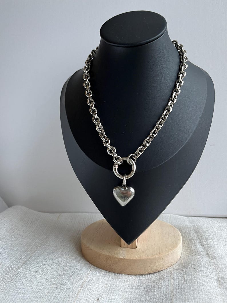 May include: A silver-toned chain necklace featuring a thick, interlocking link design. The necklace includes a circular clasp at the centre, from which a smooth, polished, heart-shaped pendant hangs. The metal has a reflective, metallic finish.