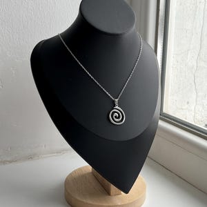 Op de afbeelding: Een zilverkleurige hangerketting met een spiraalvormig ontwerp. De hanger hangt aan een fijne ketting en wordt getoond op een zwarte sieraden buste met een houten voet. De achtergrond is neutraal.