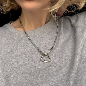 May include: Silver necklace with a heart-shaped pendant. The pendant is a solid heart design with a clasp detail. The chain is a textured, rounded link style. The necklace is worn against a grey sweatshirt.