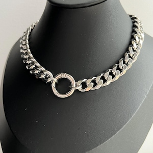 Op de afbeelding: Een zilverkleurige ketting met een groot, dik schakelontwerp. De ketting heeft een cirkelvormige sluiting in het midden. De sieraden worden getoond op een zwarte mannequinhoofd.