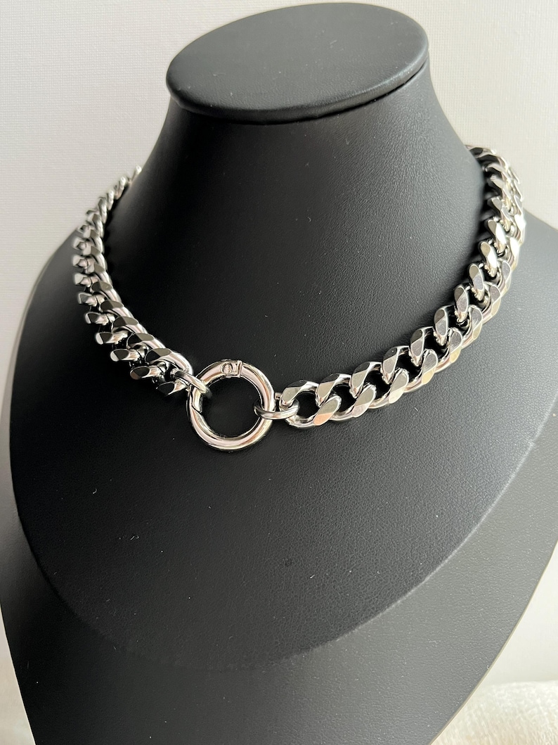 Op de afbeelding: Een zilverkleurige ketting met een groot, dik schakelontwerp. De ketting heeft een cirkelvormige sluiting in het midden. De sieraden worden getoond op een zwarte mannequinhoofd.