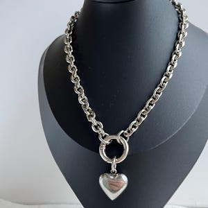 May include: A silver-toned chain necklace featuring thick, rounded links and a large circular spring-ring clasp. A smooth, polished, puffy heart-shaped pendant hangs from the centre of the clasp, creating a bold and reflective metallic accessory.