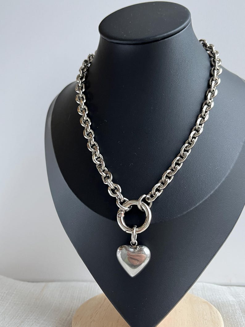May include: A silver-toned chain necklace featuring thick, rounded links and a large circular spring-ring clasp. A smooth, polished, puffy heart-shaped pendant hangs from the centre of the clasp, creating a bold and reflective metallic accessory.