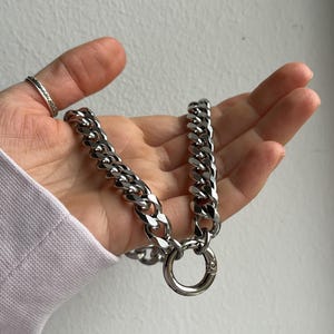 Op de afbeelding: Een zilverkleurige ketting met grote, in elkaar grijpende schakels. De ketting heeft een ronde sluiting. De sieraden worden in een hand gehouden, met een ring aan de wijsvinger. De achtergrond is een effen, gebroken witte muur.