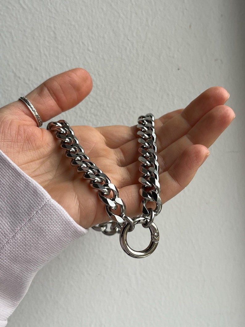 Op de afbeelding: Een zilverkleurige ketting met grote, in elkaar grijpende schakels. De ketting heeft een ronde sluiting. De sieraden worden in een hand gehouden, met een ring aan de wijsvinger. De achtergrond is een effen, gebroken witte muur.