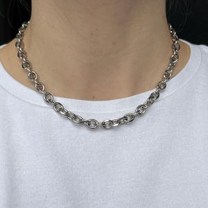 Könnte beinhalten: Eine silberfarbene Halskette mit großen ovalen Gliedern. Die Halskette wird um den Hals getragen und liegt auf einem weißen T-Shirt. Die Kette besteht aus glänzendem Metall.