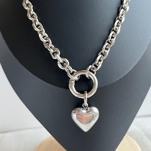 May include: A silver-toned chain necklace featuring thick, rounded links and a large circular spring-ring clasp at the centre. A smooth, polished, puffed heart-shaped pendant hangs from the clasp, creating a bold and reflective metallic accessory.
