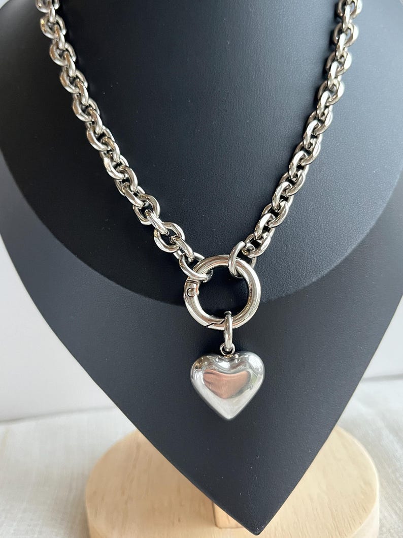 May include: A silver-toned chain necklace featuring thick, rounded links and a large circular spring-ring clasp at the centre. A smooth, polished, puffed heart-shaped pendant hangs from the clasp, creating a bold and reflective metallic accessory.