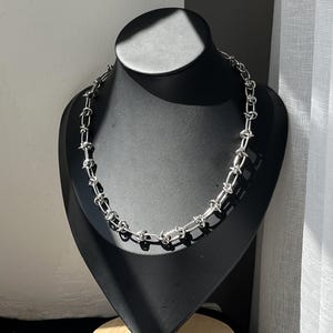 May include: A silver-toned chain necklace featuring a series of interconnected links that resemble barbed wire. The necklace is displayed on a black bust, showcasing a metallic finish with a repeating pattern of twisted wire segments throughout the chain.