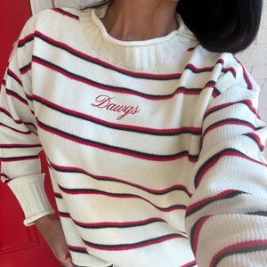 Puede incluir: Un suéter de punto color crema con rayas horizontales en rojo y negro. El suéter tiene un cuello redondo acanalado y puños acanalados. La palabra "Dawgs" está bordada en letra cursiva roja en el centro de la zona del pecho.