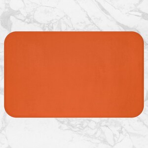 Può includere: Un tappetino da bagno rettangolare arancione con angoli arrotondati. Il tappetino è di un colore arancione solido e vibrante e sembra essere realizzato in un materiale morbido e assorbente. Lo sfondo è una superficie di marmo bianco.
