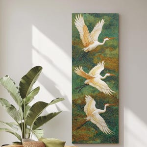 May include: A vertical canvas art piece featuring three white cranes in flight against a textured green and gold background. The birds have orange beaks and accents, with outstretched wings. The artwork is displayed in a modern living room setting.
