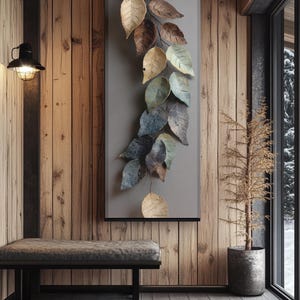 Op de afbeelding: Metalen wandkunst met een waterval van gestileerde bladeren in goud, bruin en blauwgroen. De bladeren zijn gemonteerd op een grijs rechthoekig paneel, waardoor een op de natuur ge&iuml;nspireerd decoratief element ontstaat. Het kunstwerk wordt getoond in een rustiek interieur.