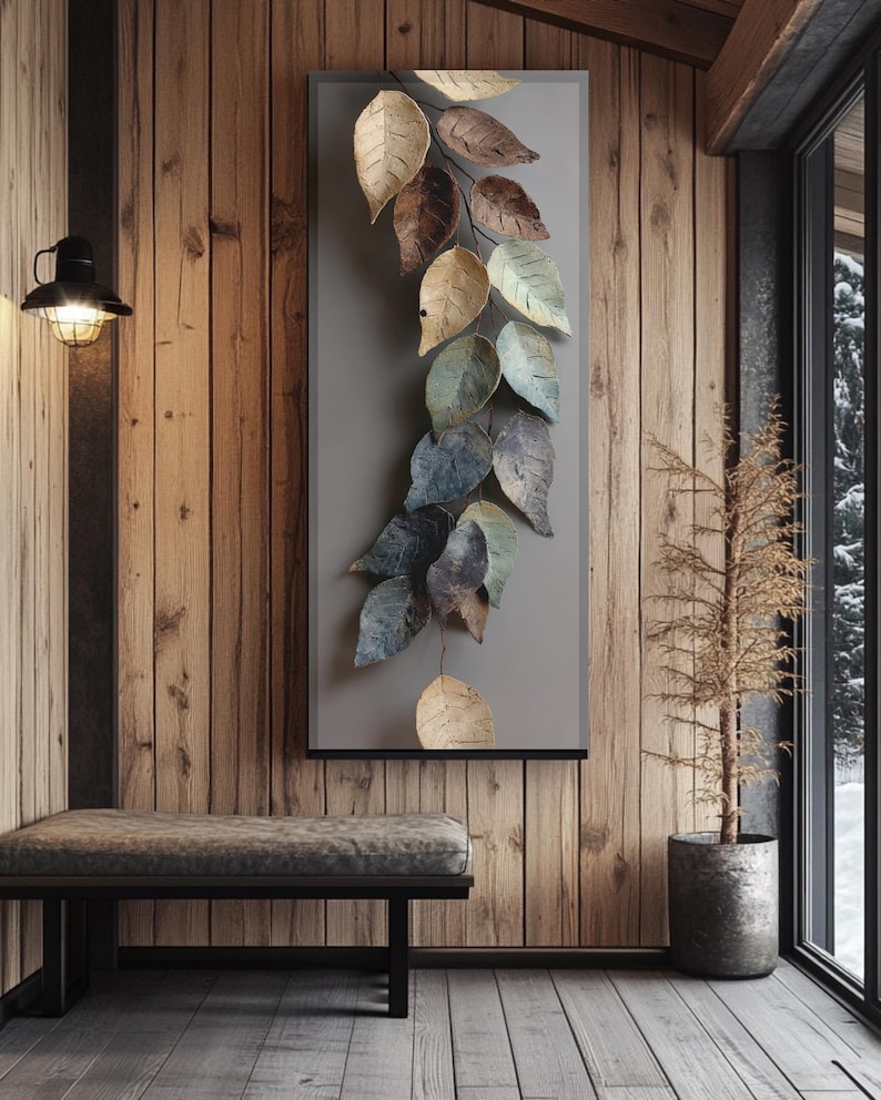 Op de afbeelding: Metalen wandkunst met een waterval van gestileerde bladeren in goud, bruin en blauwgroen. De bladeren zijn gemonteerd op een grijs rechthoekig paneel, waardoor een op de natuur ge&iuml;nspireerd decoratief element ontstaat. Het kunstwerk wordt getoond in een rustiek interieur.
