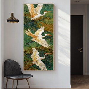 May include: Vertical framed artwork featuring three white cranes in flight against a green and gold textured background. The cranes have orange beaks and wing tips. The artwork is displayed on a white wall next to a black chair.