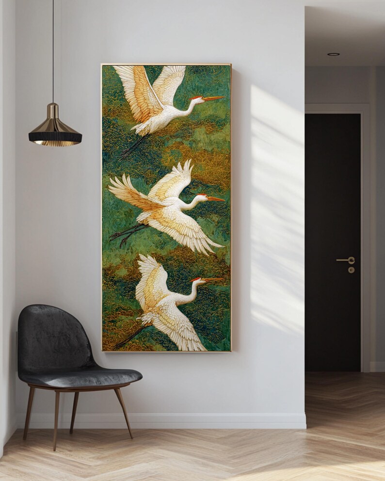 May include: Vertical framed artwork featuring three white cranes in flight against a green and gold textured background. The cranes have orange beaks and wing tips. The artwork is displayed on a white wall next to a black chair.