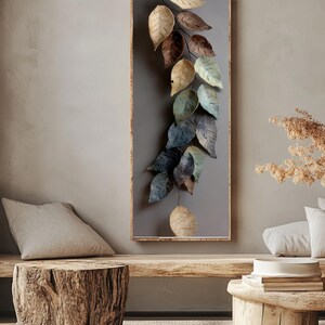 Op de afbeelding: Een rechthoekig wandkunstwerk met een houten frame, met een rank van gestileerde bladeren in goud, bruin en blauwgroen. De bladeren zijn geplaatst tegen een grijze achtergrond, wat een natuurlijke esthetiek cre&euml;ert. Het kunstwerk wordt tentoongesteld in een neutraal getinte woonruimte.
