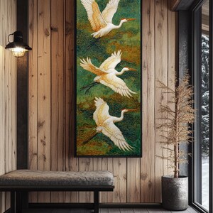 May include: A framed artwork featuring three white and gold cranes in flight against a green and gold textured background. The birds have outstretched wings and long legs. The artwork is displayed on a wooden wall in a room with a bench and a window.