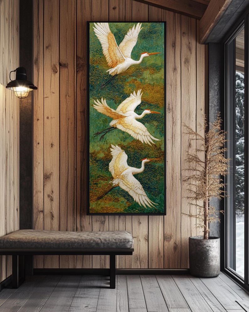 May include: A framed artwork featuring three white and gold cranes in flight against a green and gold textured background. The birds have outstretched wings and long legs. The artwork is displayed on a wooden wall in a room with a bench and a window.