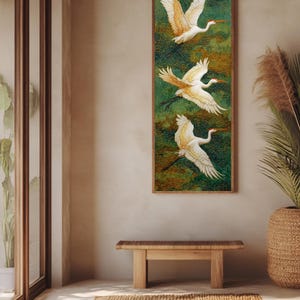 May include: A vertical framed artwork features three white birds with outstretched wings against a green and gold background. The birds have orange beaks and accents. The artwork hangs on a neutral-colored wall, above a wooden bench and a woven rug.