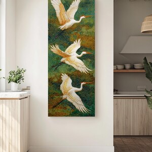 May include: A vertical canvas art piece featuring three white cranes in flight against a textured green and gold background. The cranes have outstretched wings with detailed feathering and orange beaks, creating a sense of movement and elegance. The artwork is displayed in a well-lit room.