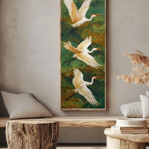 May include: A vertical rectangular artwork features three white cranes in flight against a green and gold background. The birds have outstretched wings and long legs. The artwork is framed in a light brown wooden frame, creating a serene and natural aesthetic.