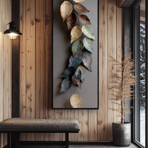 Op de afbeelding: Een ingelijst wandkunstwerk met een metalen bladontwerp in goud, bruin en blauwgroen. Het kunstwerk is gemonteerd op een grijze achtergrond en zwart omlijst. Het hangt aan een houten muur.