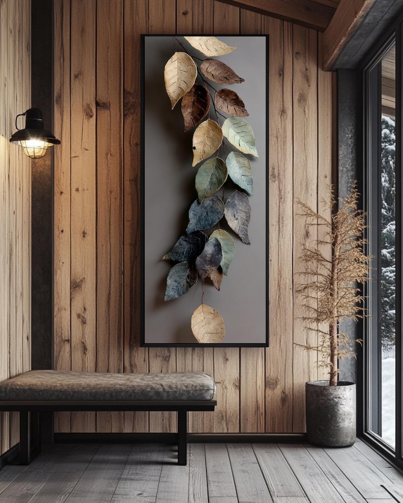 Op de afbeelding: Een ingelijst wandkunstwerk met een metalen bladontwerp in goud, bruin en blauwgroen. Het kunstwerk is gemonteerd op een grijze achtergrond en zwart omlijst. Het hangt aan een houten muur.