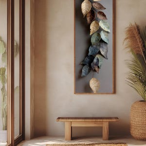 Op de afbeelding: Een verticaal wandkunstwerk met een waterval van metalen bladeren in tinten beige, bruin en blauwgroen. De bladeren zijn gemonteerd op een grijze achtergrond in een houten frame. Een houten bank en een geweven vloerkleed staan op de voorgrond.