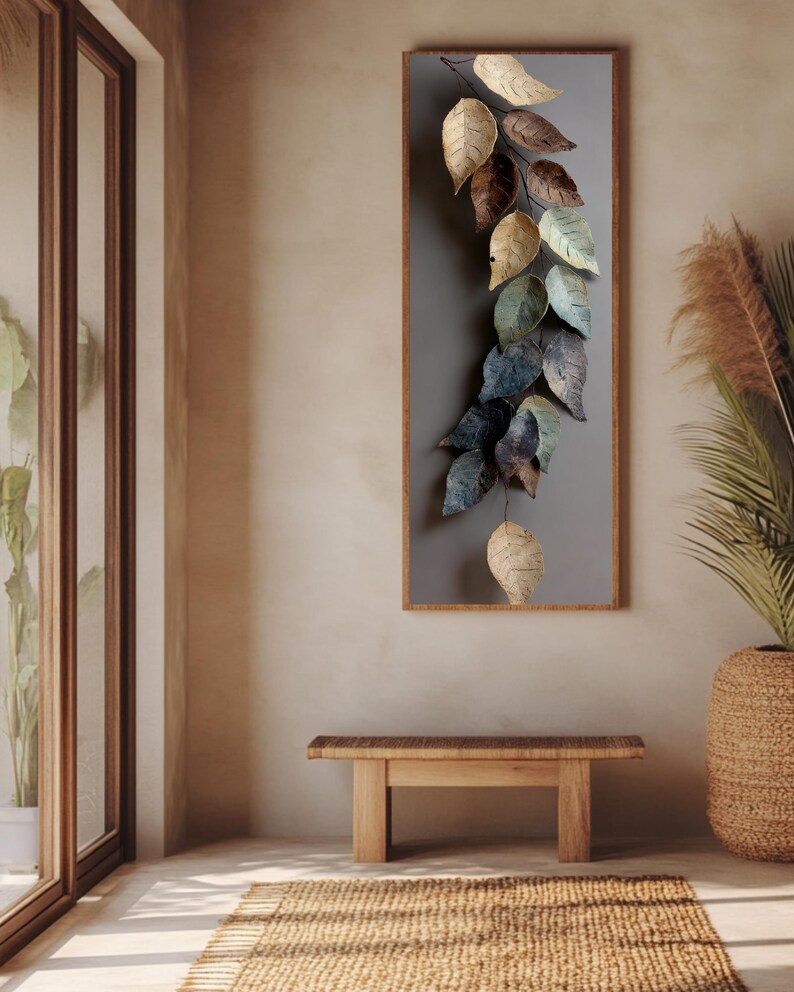 Op de afbeelding: Een verticaal wandkunstwerk met een waterval van metalen bladeren in tinten beige, bruin en blauwgroen. De bladeren zijn gemonteerd op een grijze achtergrond in een houten frame. Een houten bank en een geweven vloerkleed staan op de voorgrond.