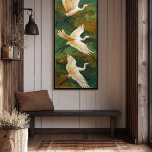 May include: A vertical framed artwork features three white birds with orange beaks and wingtips in flight against a green and gold background. The artwork hangs above a dark wooden bench in a rustic interior setting.