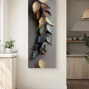 Op de afbeelding: Een verticaal kunstwerk voor aan de muur met een tak met meerdere bladeren in tinten beige, bruin en blauwgroen. De bladeren hebben een textuur en zijn geplaatst tegen een grijze achtergrond, wat een natuurlijk, decoratief element cre&euml;ert.