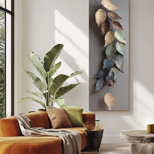 Op de afbeelding: Een moderne woonkamer met een oranje hoekbank, een grote plant in een pot en een houten bijzettafel. Een decoratieve wanddecoratie toont metalen bladeren in tinten bruin, goud en blauw. De kamer heeft een neutraal kleurenpalet.