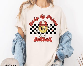 May include: Ivory-colored t-shirt featuring a softball graphic. The graphic includes a cartoon softball character wearing a red cap, waving, and standing in front of a black and white checkered pattern. The text reads "Lady 5g Prime Softball" in red.